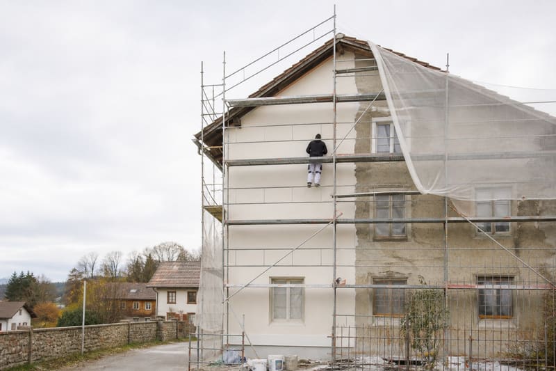 Hausfassade unter Gerüst und Schutznetz, ein Arbeiter auf dem Gerüstboden